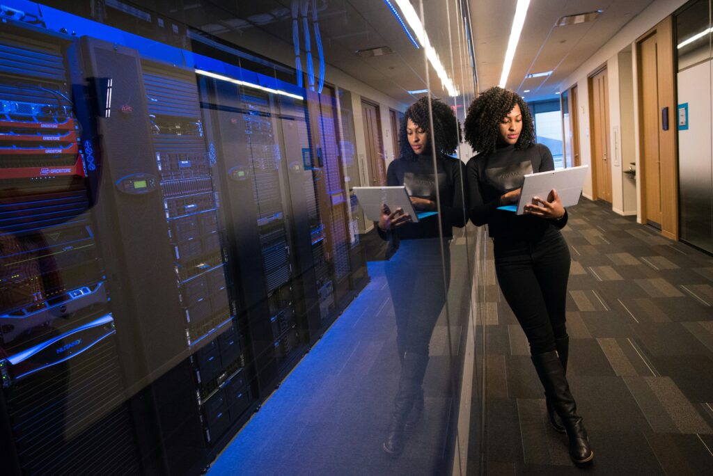 Women with dark hair stoof in a corridor holding a laptop with a computer server room to her left.