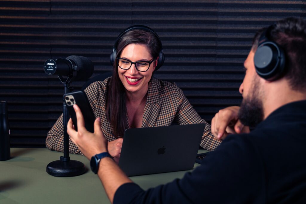 woman with borwn hair facing the camera with a podcast icrophone in front of her. She is sat opposite a man in headphones with his back to the camera