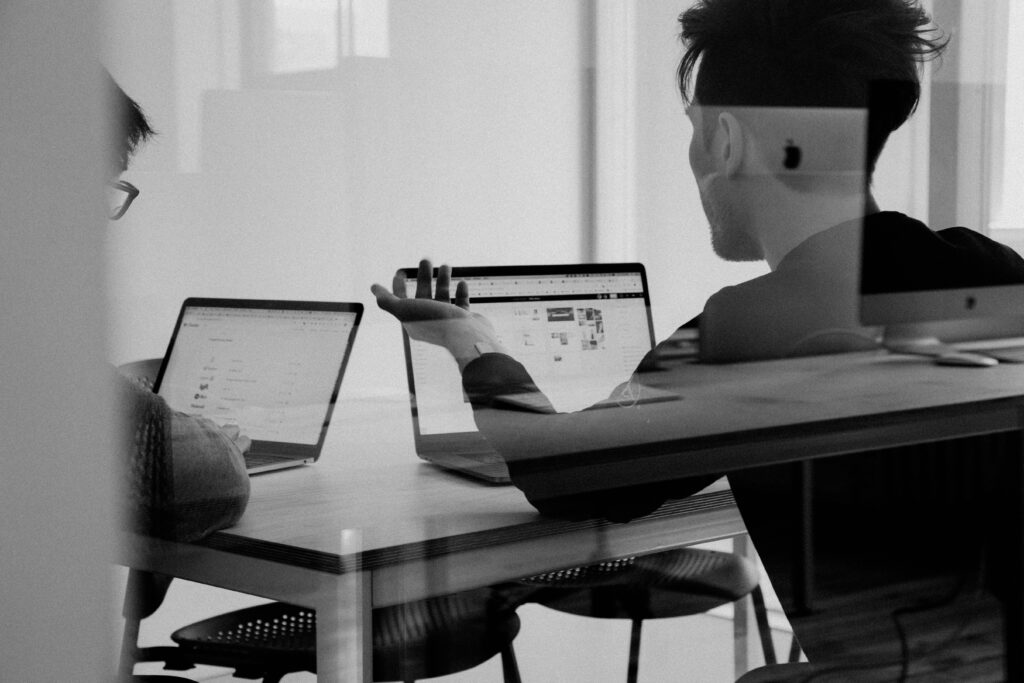 Black and white image of two people sat in an office talking over mac books.