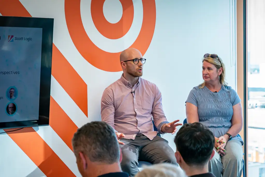 Two people as part of a panel, with one of them speaking to an audience in front of them.