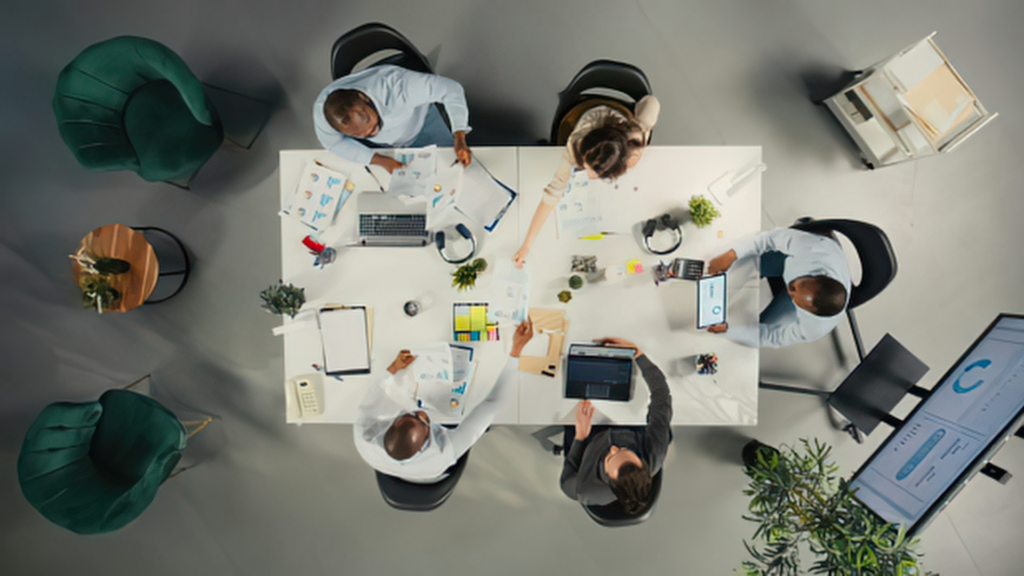 Professional group collaborating in a bright, modern workspace with laptops, papers and plants around a large rectangular table.